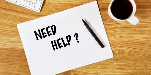 A white sheet of paper on a wooden desk with the words 'NEED HELP' written on it in black ink.